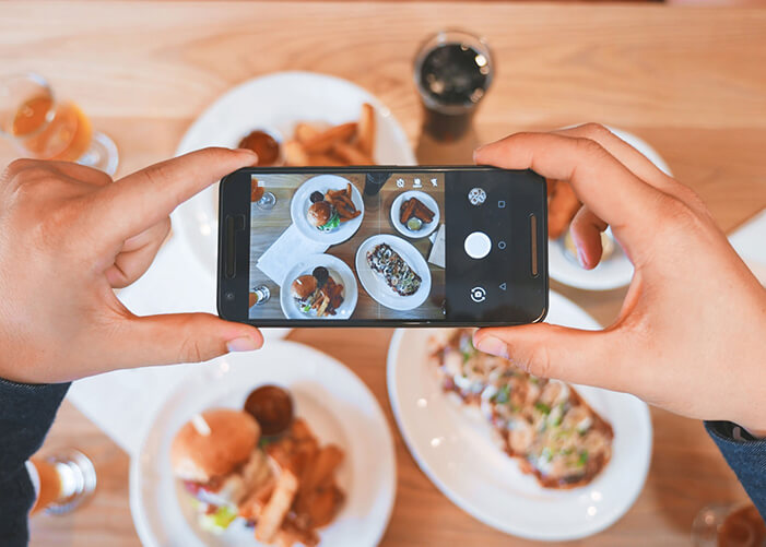 Mãos segurando celular, fotografando 4 pratos com comidas diferentes